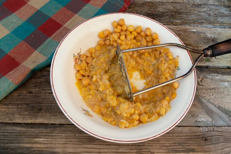 Cooked chickpeas in a white bowl being mashed with a metal potato masher.