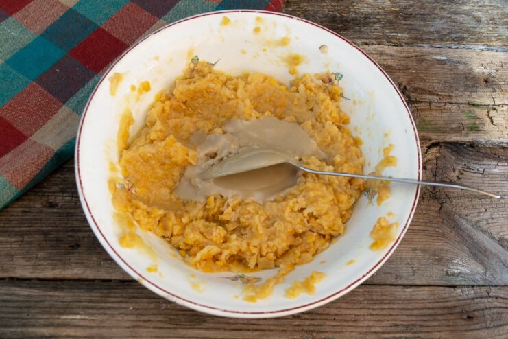 Tahini paste being added from a spoon into a bowl of mashed chickpeas.