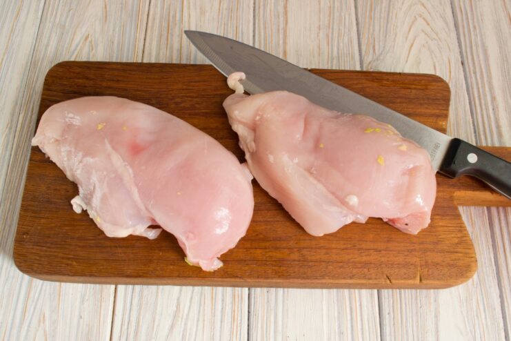 How to Prepare Chicken Breast for Fricassee A hand holding a knife is cutting a raw chicken breast off the bone on a cutting board.