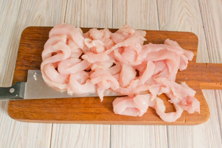 Cutting Chicken into Strips Against the Grain Close-up of raw chicken fillet being sliced into thin strips on a cutting board.