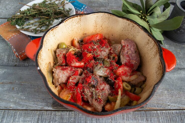 Seasoning the stew with paprika, oregano, salt, and pepper. A hand sprinkling a mix of spices into the braiser with beef and vegetables.