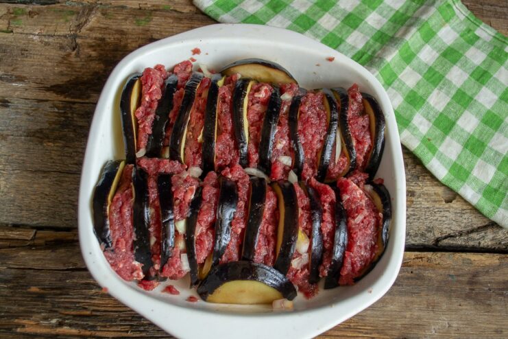 Layering Eggplant and Beef in the Baking Dish Eggplant slices topped with ground beef arranged tightly in a ceramic baking dish.