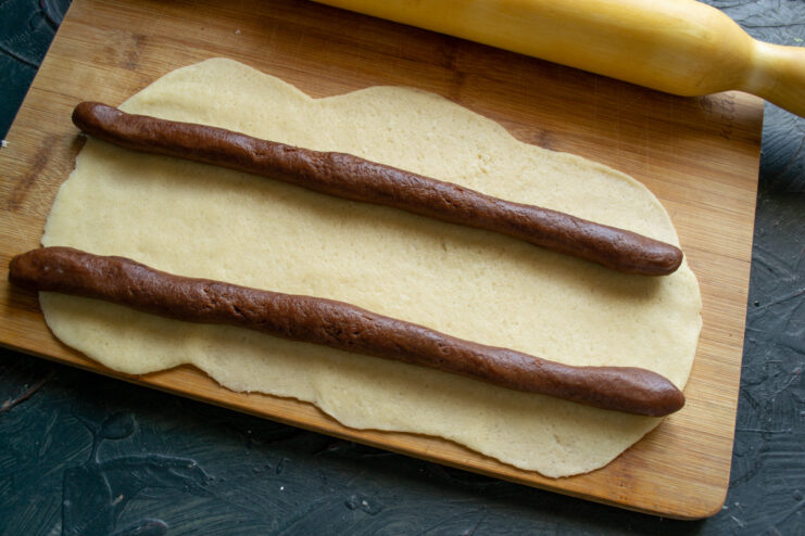 Placing chocolate dough logs on the plain dough sheet Two logs of chocolate dough placed parallel on top of the rolled-out plain dough.