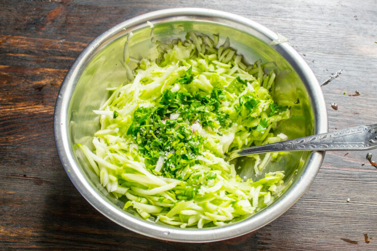 Adding Fresh Herbs to the Zucchini Mixture A bowl of grated zucchini with freshly chopped parsley being added.