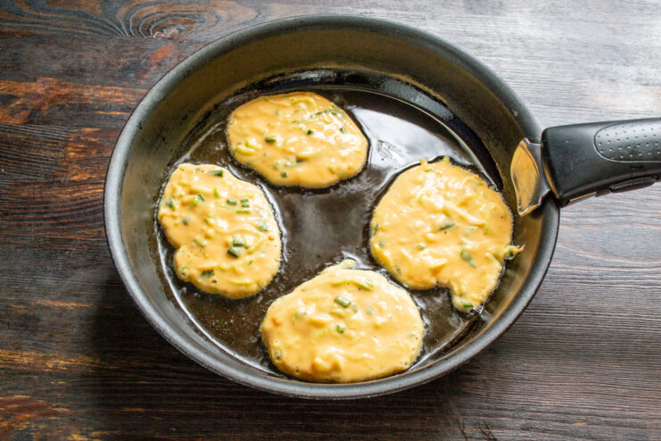 Frying Zucchini Fritters in a Hot Pan Spoonfuls of zucchini fritter batter being placed into a hot frying pan with oil.