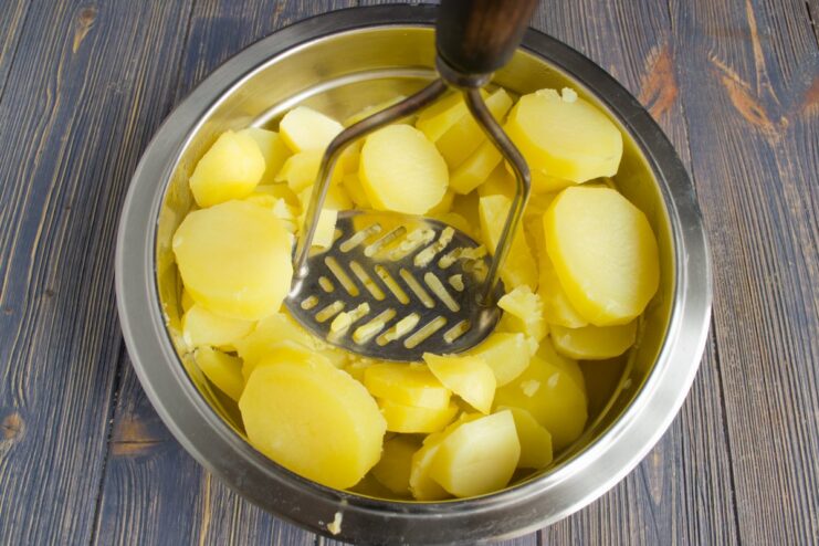 Mashing Boiled Potatoes for a Smooth Casserole Topping A metal potato masher mashing boiled potatoes in a white bowl to create a smooth puree.
