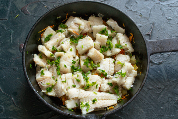 Layering the deboned fish over the cooked vegetables Flaked, cooked fish being layered over a bed of sautéed carrots and onions in a skillet.