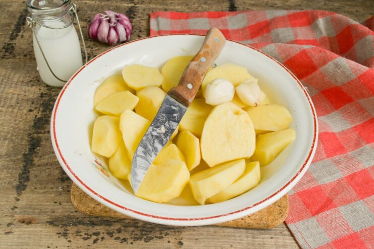 Thickly sliced raw potatoes and peeled garlic cloves on a cutting board.