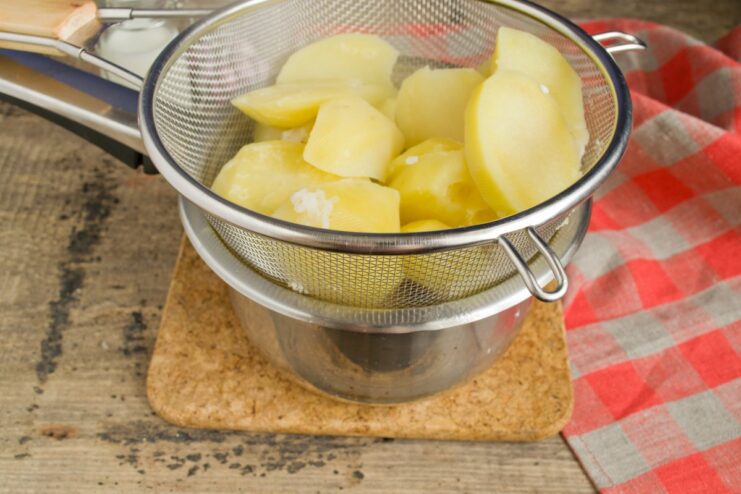 Cooked potato slices being drained in a metal sieve over a sink.