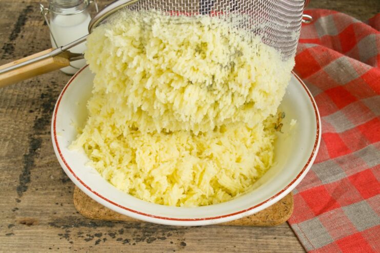 A hand using a spoon to press cooked potatoes through a fine-mesh sieve into a bowl.