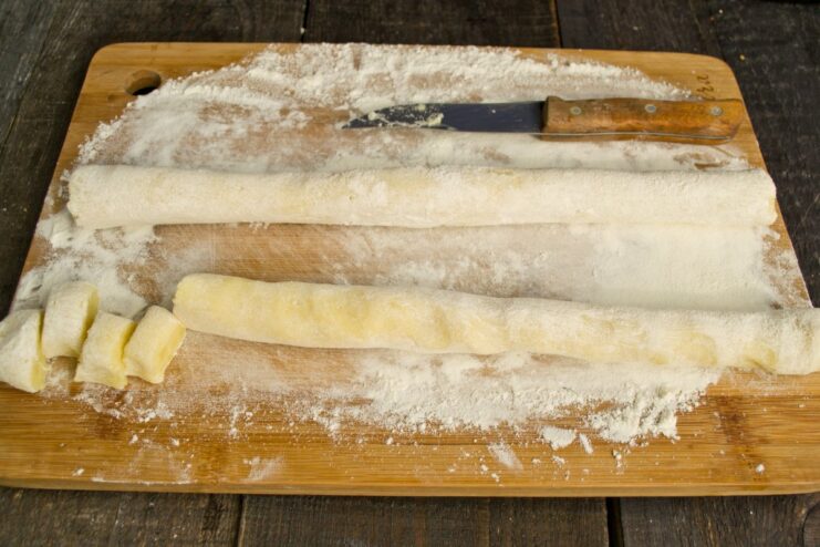 Shaping and Cutting the Švilpikai Dumplings Potato dough rolled into a log on a floured surface, being cut into small, round dumpling pieces.