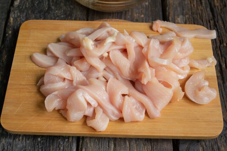 Raw chicken fillet being sliced into thin strips on a cutting board.