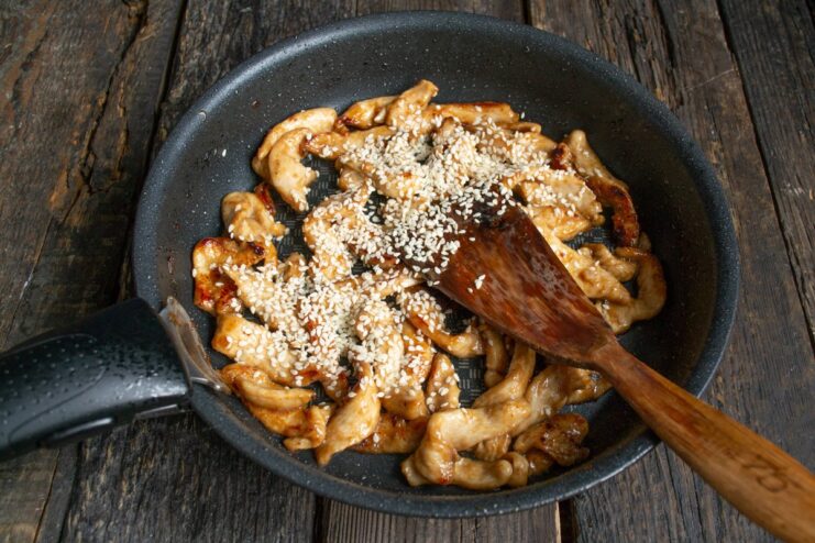 Sprinkling white sesame seeds over the cooking teriyaki chicken in the pan.
