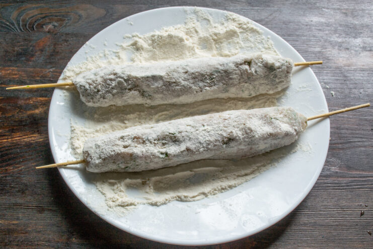 First Step of Breading: Coating in Flour A sausage on a skewer being rolled in flour on a wooden board.