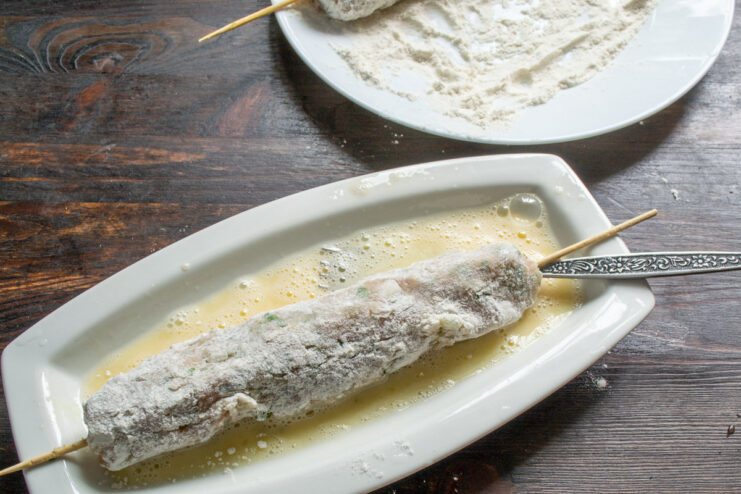 Second Step of Breading: Egg Wash A flour-coated sausage on a skewer being dipped into a narrow dish with beaten egg.