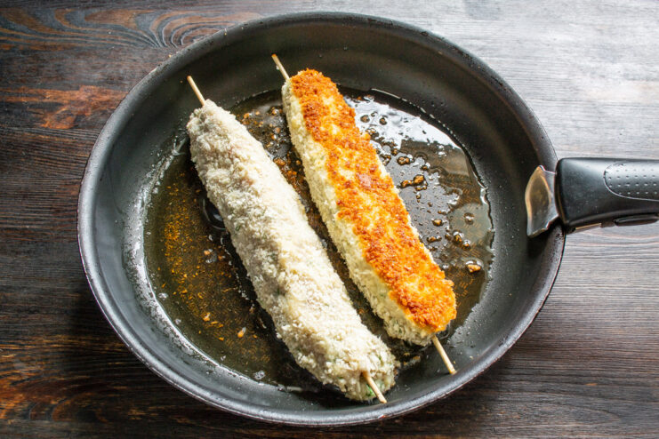 Frying the Sausages to a Golden Crisp Two breaded sausages on skewers frying in a pan with hot oil, turning golden brown.