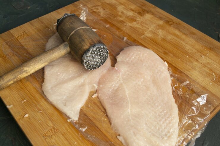Step 2: Pounding the chicken fillet to make it thin and even Chicken fillet covered with plastic wrap being pounded with a meat mallet.