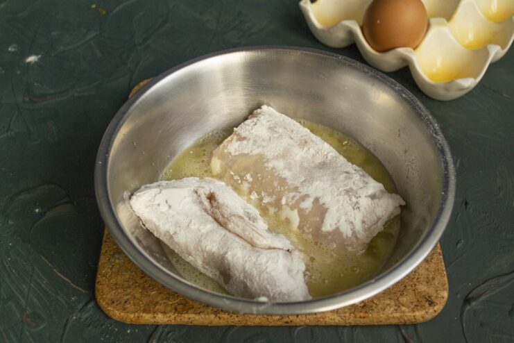 Step 5: Dipping the floured cutlet into the egg wash A flour-coated chicken cutlet being dipped into a bowl of beaten eggs.