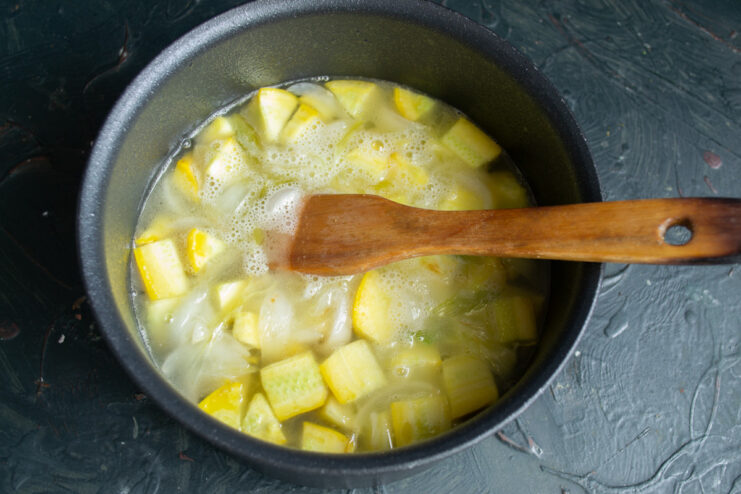 Adding liquid: pour in broth to cook the vegetables Broth being poured into a pot with sautéed onions and diced zucchini.