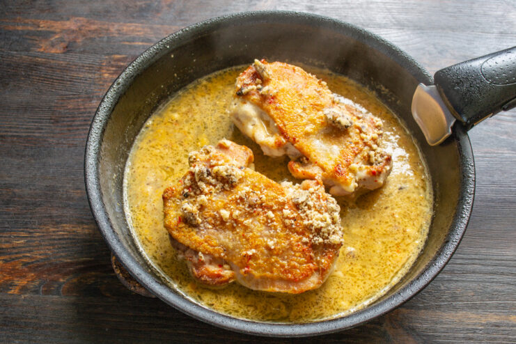 Adding Walnuts and Ginger Sauce to the Seared Chicken Seared chicken thighs in a skillet, being topped with crushed walnuts and a liquid ginger sauce.