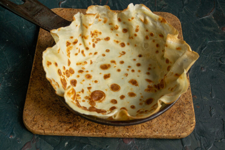 The first pancake being laid inside a greased baking dish, with edges hanging over.