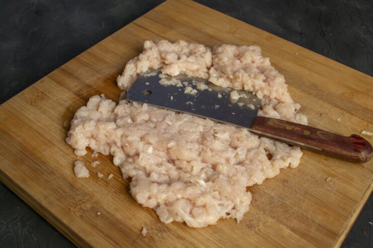 Preparing the chicken fillet by dicing it into small cubes for the filling. Raw chicken breast fillet being cut into small cubes on a cutting board.