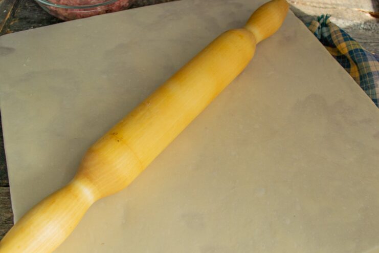 Rolling the Dough Thinly for Lazy Pelmeni A large, thin sheet of dough rolled out on a wooden board with a rolling pin.