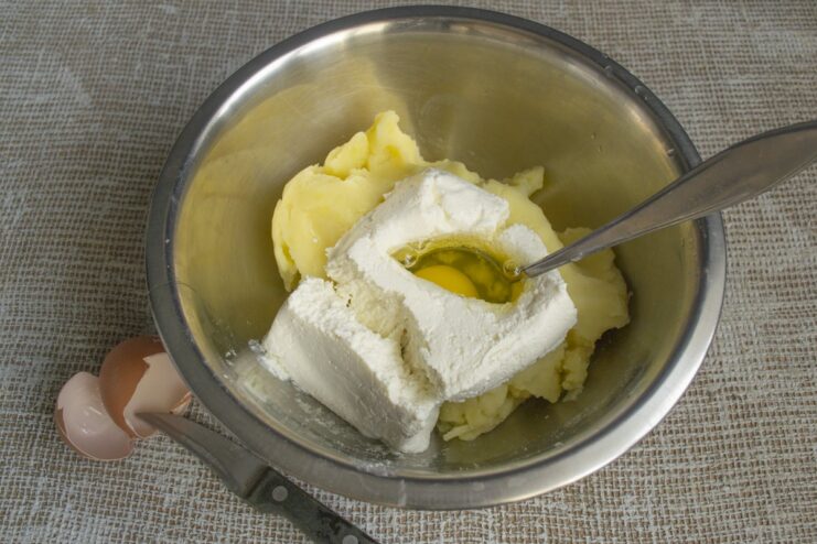 A raw egg being added to the potato and tvorog mixture in a bowl.