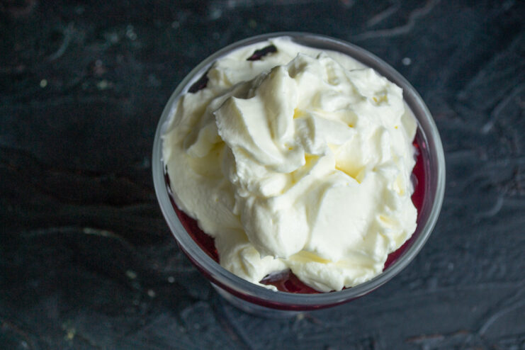 Adding a generous layer of whipped cream Spoonfuls of freshly whipped cream being placed on top of set raspberry jelly in a glass.