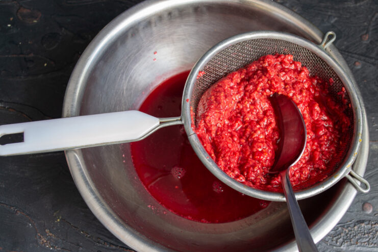 How to get a smooth, seedless raspberry puree A hand using a spoon to press cooked raspberries through a fine-mesh sieve over a bowl.