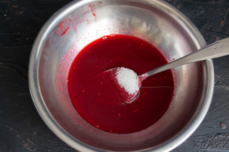 Sweetening the raspberry juice for the jelly Strained raspberry juice in a glass bowl with granulated sugar being added.