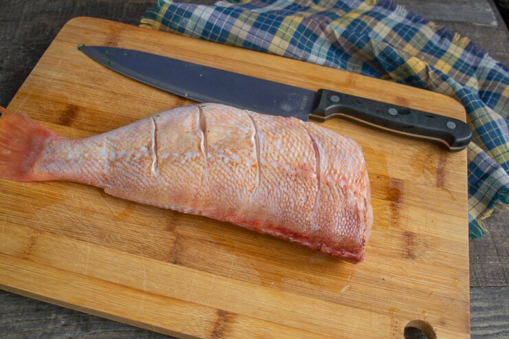 A whole sea bass with diagonal scores on its side, being seasoned with sea salt.