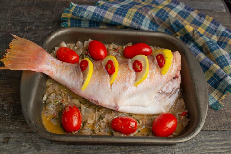 Sea bass in a baking dish, stuffed with lemon slices and chili rings, surrounded by cherry tomatoes.