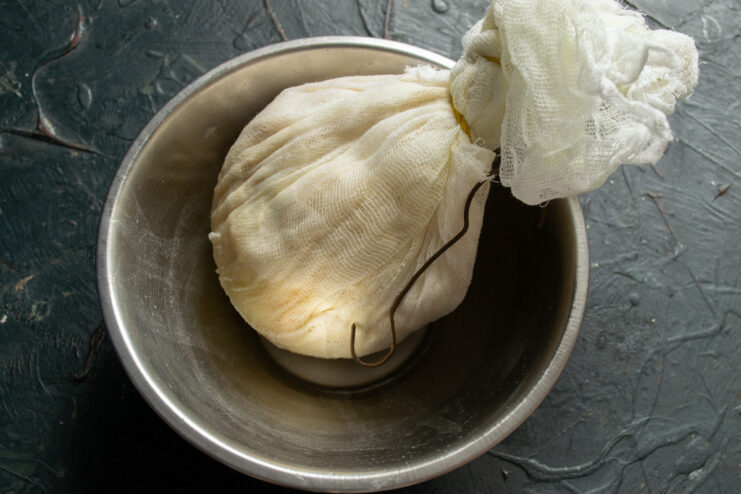 Draining the Cheese — The longer it drains, the firmer the cheese will be. A cheesecloth bundle hanging to drain whey into a bowl placed underneath.