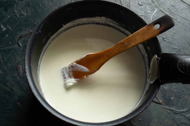 Heating the Smetana and Milk Mixture — Heat slowly and stir constantly to avoid boiling. A saucepan on a stove with a white dairy mixture being heated, showing steam rising.