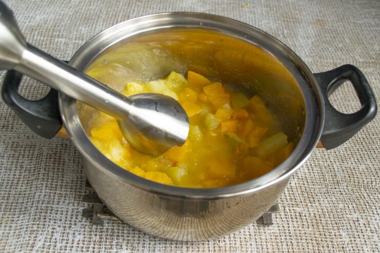 Using an immersion blender to create a smooth, creamy purée. An immersion blender pureeing the cooked pumpkin mixture directly in the pot.