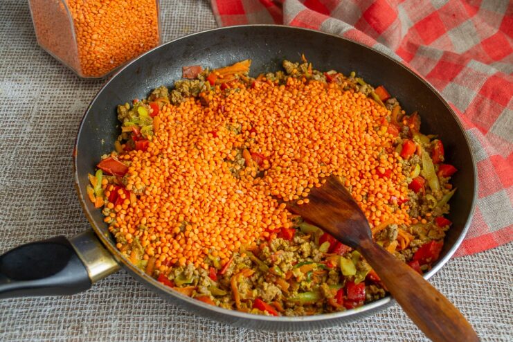 Pouring dry red lentils into the simmering chili con carne. Dry red lentils being poured into the skillet with the chili con carne.