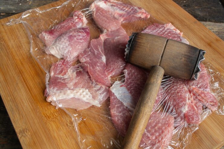 Step 2: Pounding the Pork Cutlets Pork cutlets covered in plastic wrap being pounded with a meat mallet.