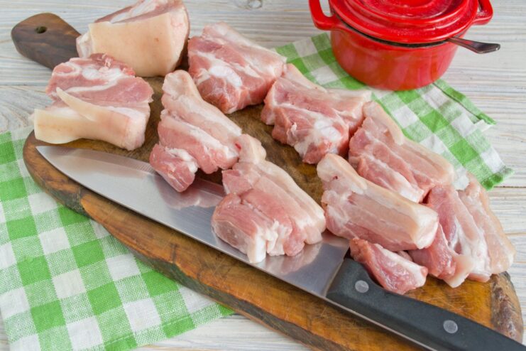 Cut the pork belly into thick slices for even roasting. Raw pork belly being cut into thick slices on a cutting board.