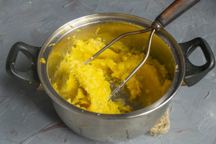 Step 6: Mashing the vegetables into a coarse puree A hand holding a potato masher, mashing the cooked vegetables and onions in a pot.