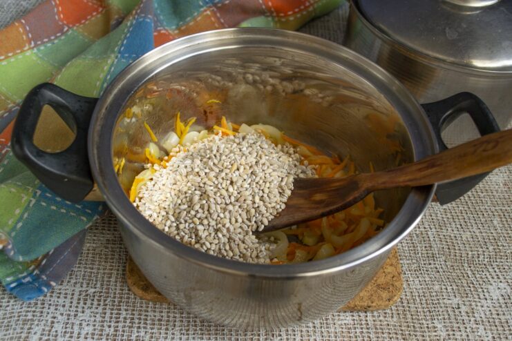 Adding pearl barley to the sautéed vegetables. Dry pearl barley being added to the pot of cooked vegetables.