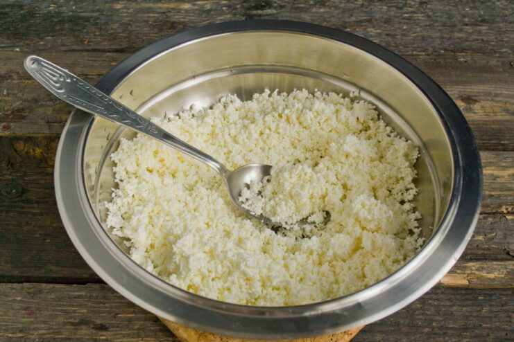 Preparing the Creamy Filling: Sifting Tvorog for a Smooth Texture Hands sifting white farmer's cheese through a fine-mesh sieve into a bowl.