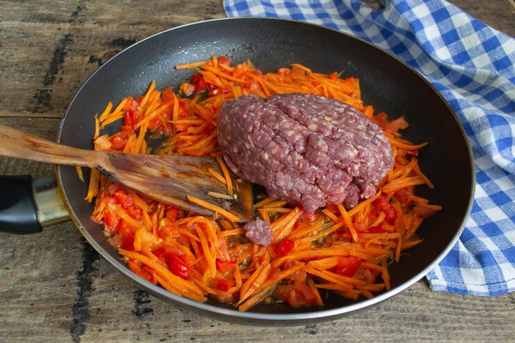 Ground beef being added to a pan of sautéed carrots, peppers, and tomatoes.