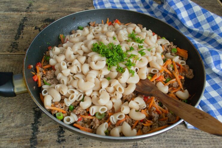 Cooked spelt pasta and fresh dill being added to the beef and vegetable sauce in a pan.