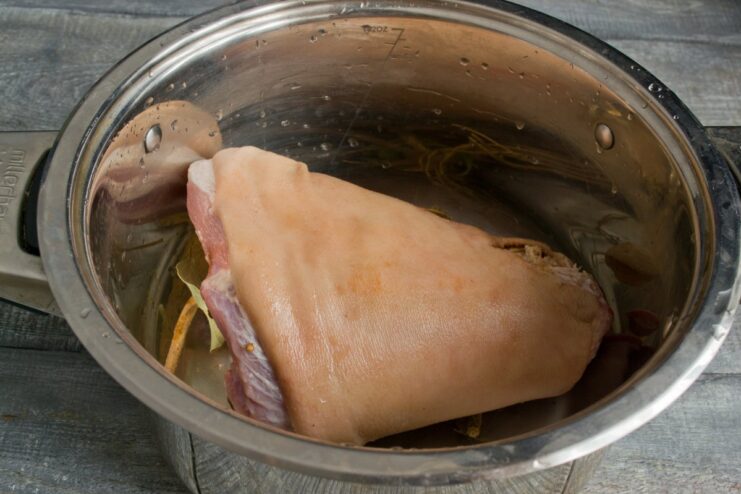 Step 2: Placing the Pork Knuckle into the Pot for Boiling A raw pork knuckle being placed into a pot on top of a bed of spices.