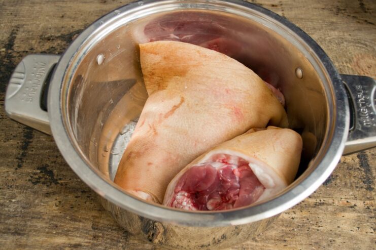 Step 1: Placing the Pork Knuckles in a Large Pot to Start Cooking Two raw pork knuckles being placed inside a large metal cooking pot.