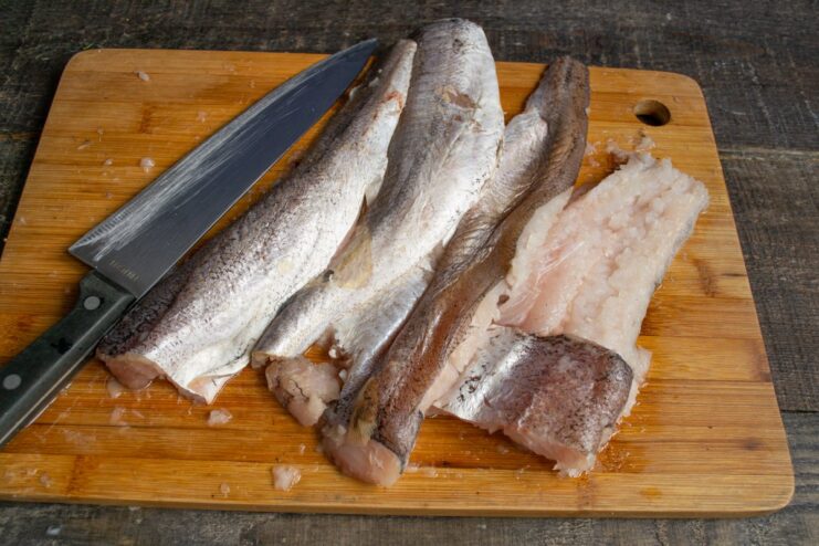 Preparing the hake fillet by removing the bones A hand deboning a thawed hake fillet on a cutting board.