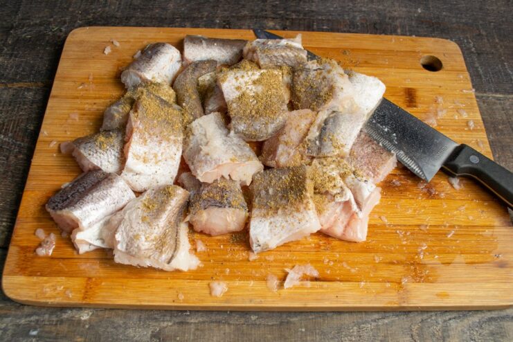 Seasoning the cut pieces of hake fillet before cooking Pieces of raw hake fillet seasoned with spices on a cutting board.