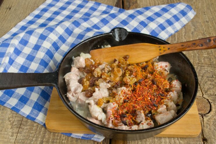Adding dried fruit and spices to the meat filling Meat mixture in a pan with raisins, barberries, and spices being added.