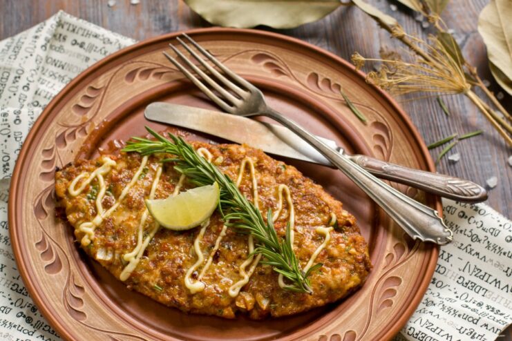 A plate with a golden-brown baked chopped chicken schnitzel, garnished with a sprig of rosemary and a lemon wedge.